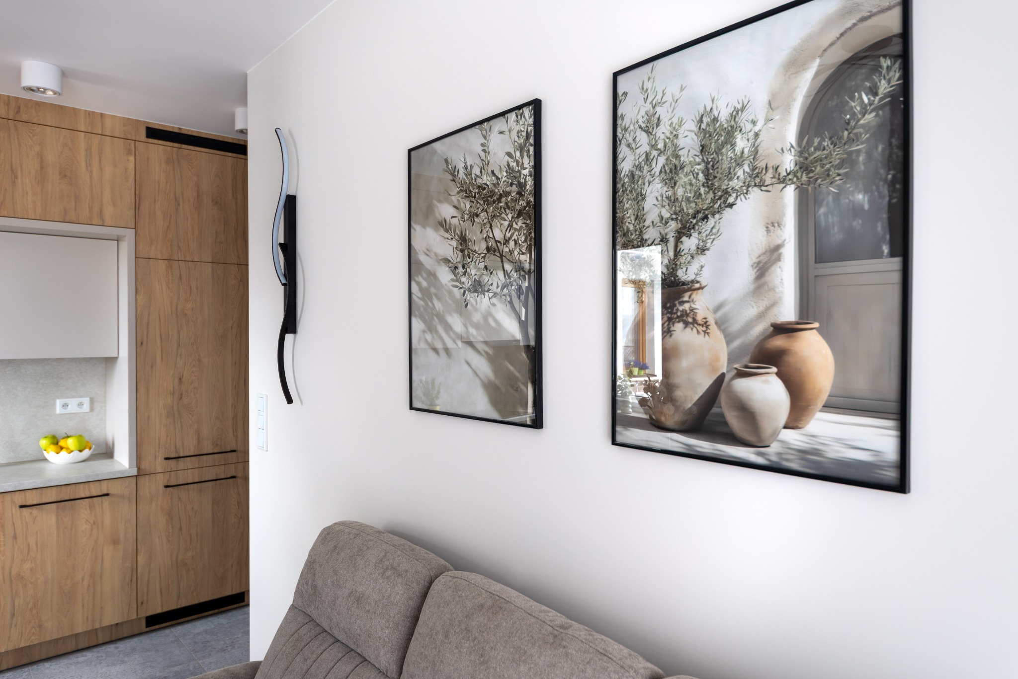 Nowoczesne wnętrze: drewniana zabudowa kuchenna, szara sofa i dekoracyjne obrazy z motywem roślinnym w glinianych wazonach na białej ścianie. Minimalistyczny design.