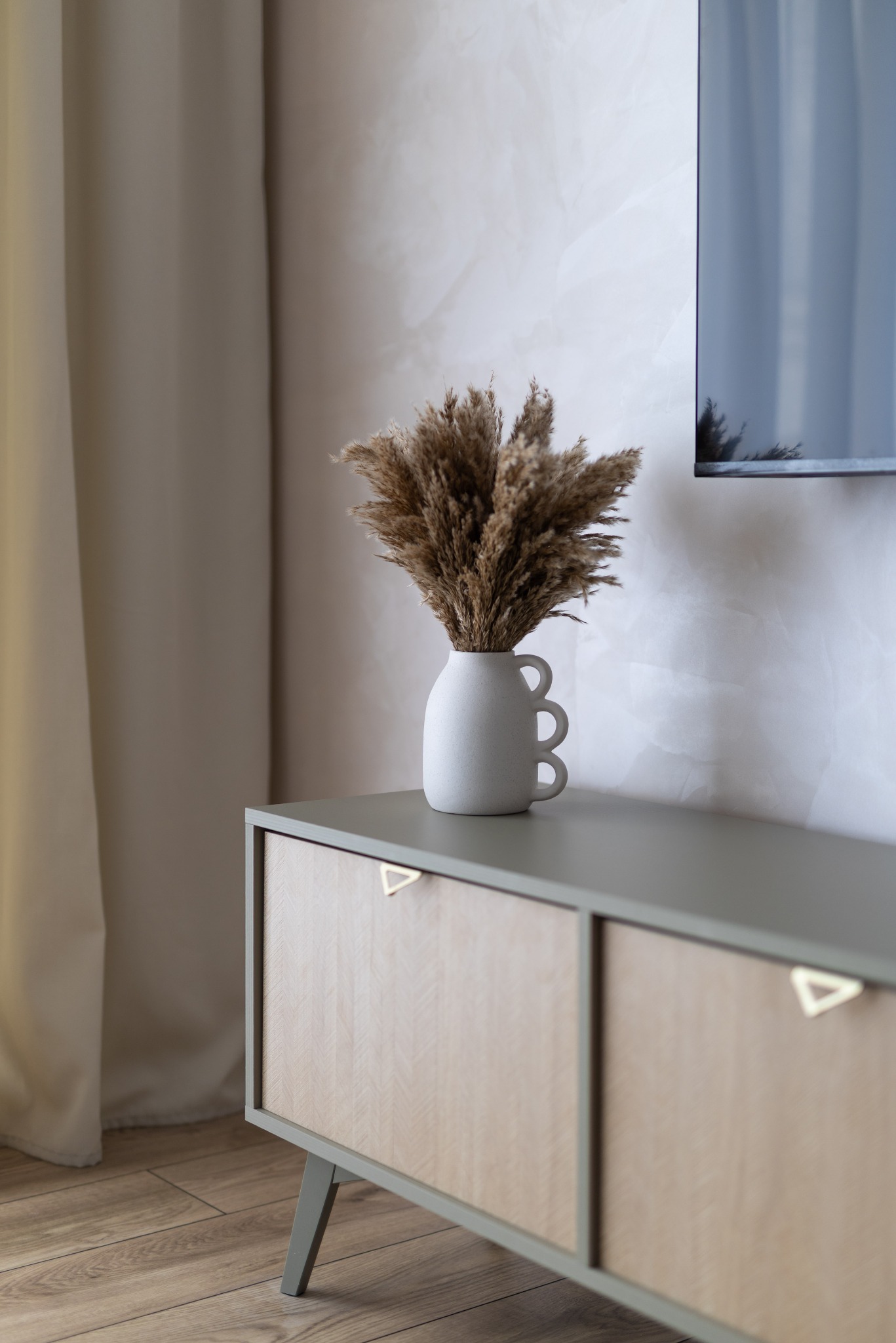 Stylowa komoda z drewnianymi frontami i trójkątnymi uchwytami, na której stoi wazon z suszonymi trawami. W tle ściana z dekoracyjnym tynkiem i fragment telewizora.
