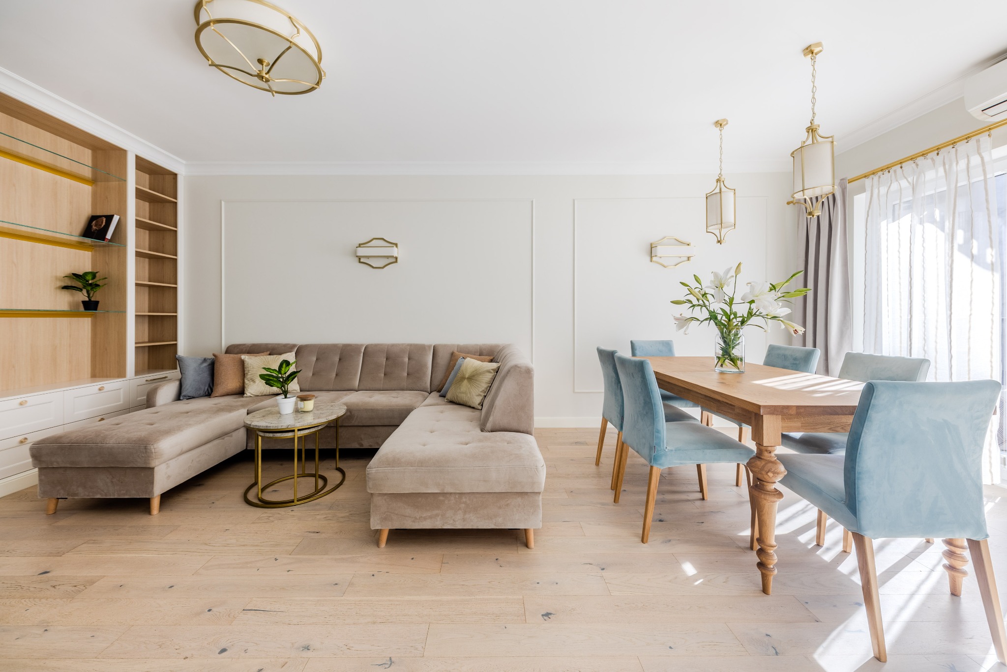 Jasne wnętrze: beżowy narożnik, drewniany stół z błękitnymi krzesłami, regał z półkami. Całość doświetlona naturalnym światłem wpadającym przez okno.