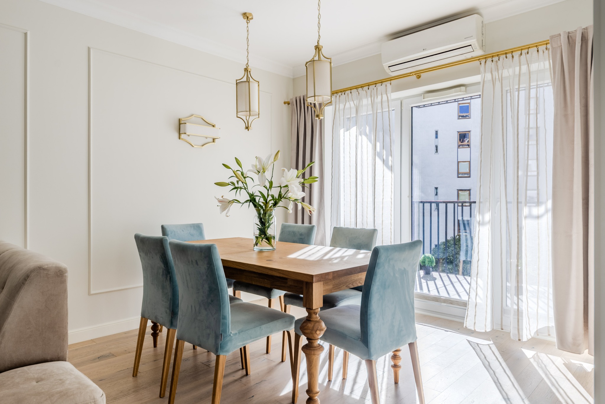 Jasna jadalnia z drewnianym stołem, sześcioma krzesłami w odcieniach błękitu i wazonem z białymi liliami. Widok na balkon przez przeszklone drzwi.