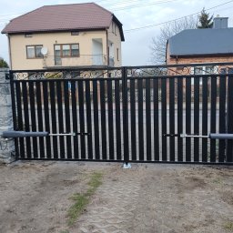 SPEEDLUK Łukasz Nowak - Automatyczna brama wjazdowa z czarnymi sztachetami i kamiennymi słupkami. Widoczne siłowniki i kostka brukowa na podjeździe. Tło stanowią dwa domy.
