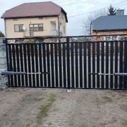 Automatyczna brama wjazdowa z czarnymi sztachetami i kamiennymi słupkami. Widoczne siłowniki i kostka brukowa na podjeździe. Tło stanowią dwa domy.