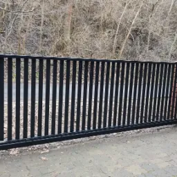 Nowoczesna, przesuwna brama wjazdowa z automatyką, czarne pionowe sztachety, murowane słupki z cegły, widoczny napęd. Tło: zieleń i drzewa bezlistne.