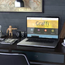 Laptop wyświetlający stronę internetową 'Graffi' z napisem 'Profesjonalne strony internetowe', ustawiony na biurku z drewnianą figurką, lampką i kalendarzem wskazującym dzień 05.