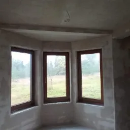 Wnętrze pokoju z trzema oknami w wykuszu, ściany pokryte świeżym, jasnoszarym tynkiem przed malowaniem.