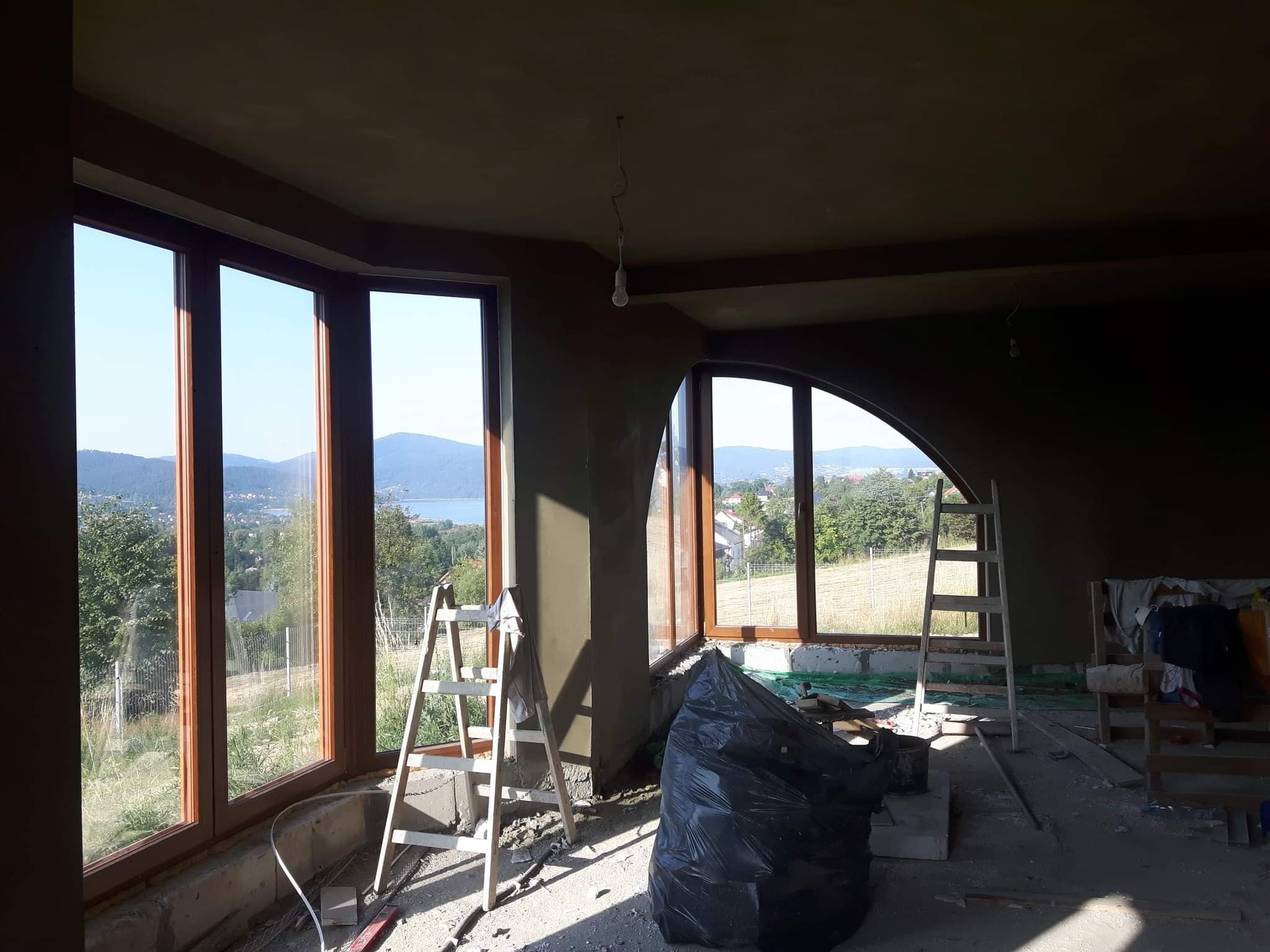 Wnętrze domu w trakcie prac wykończeniowych: świeżo nałożone tynki na ścianach, duże okna z widokiem na jezioro i góry, drabiny i worki z odpadami budowlanymi.