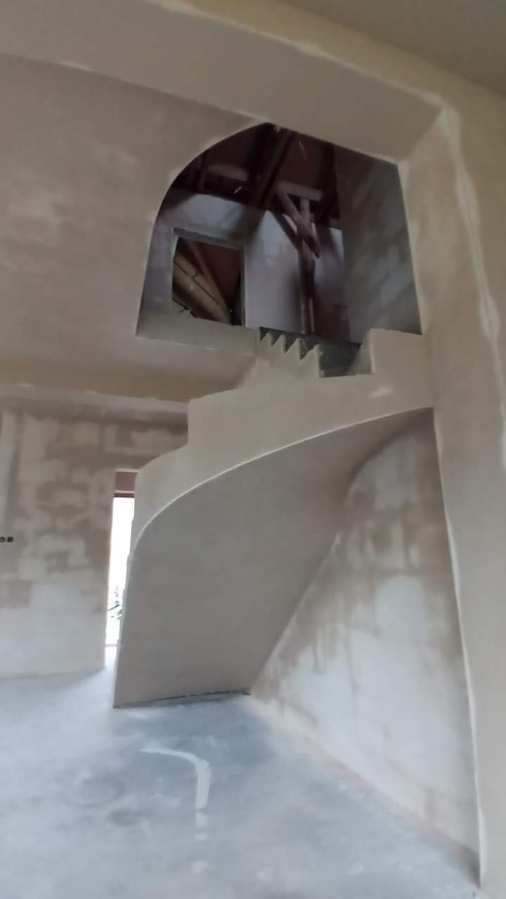 Wnętrze domu w trakcie prac wykończeniowych: zakrzywione schody z surowego tynku, ściany w trakcie gładzenia, widoczne elementy konstrukcji dachu przez otwór w ścianie.