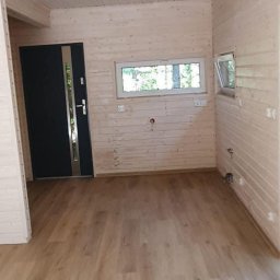 Maciej Czarnecki - Wnętrze drewnianego domku z jasnymi panelami podłogowymi, drewnianą boazerią ścienną i sufitową, czarnymi drzwiami wejściowymi z metalowymi elementami i dwoma oknami z widokiem na zieleń.