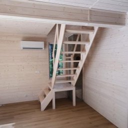 Maciej Czarnecki - Wnętrze drewnianego domu z jasną podłogą, ścianami i sufitem wykończonymi boazerią; drewniane schody ażurowe prowadzą na antresolę, na ścianie widoczny klimatyzator.