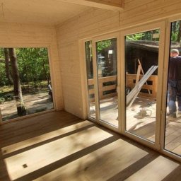 Maciej Czarnecki - Wnętrze drewnianego domku letniskowego z widokiem na las przez duże okna przesuwne, jasne drewno na ścianach, suficie i podłodze, promienie słoneczne wpadające do środka.