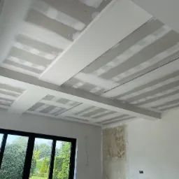 Sufit podwieszany ze szpachlowanymi łączeniami płyt w trakcie prac wykończeniowych w Zbytkowie, widok na duże okno z ogrodem w tle.