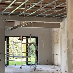 Maciej Czarnecki - Wnętrze w trakcie remontu: widoczna konstrukcja sufitu podwieszanego z instalacją elektryczną, rusztowanie przy oknie z widokiem na ogród, narzędzia i materiały budowlane na podłodze.