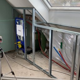 Maciej Czarnecki - Stelaż metalowy pod zabudowę w łazience na poddaszu ze skośnym oknem dachowym, widoczna instalacja wodno-kanalizacyjna Geberit i narzędzia do montażu na podłodze.