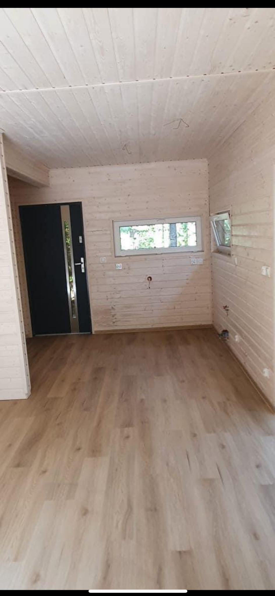 Wnętrze drewnianego domku z jasnymi panelami podłogowymi, drewnianą boazerią ścienną i sufitową, czarnymi drzwiami wejściowymi z metalowymi elementami i dwoma oknami z widokiem na zieleń.