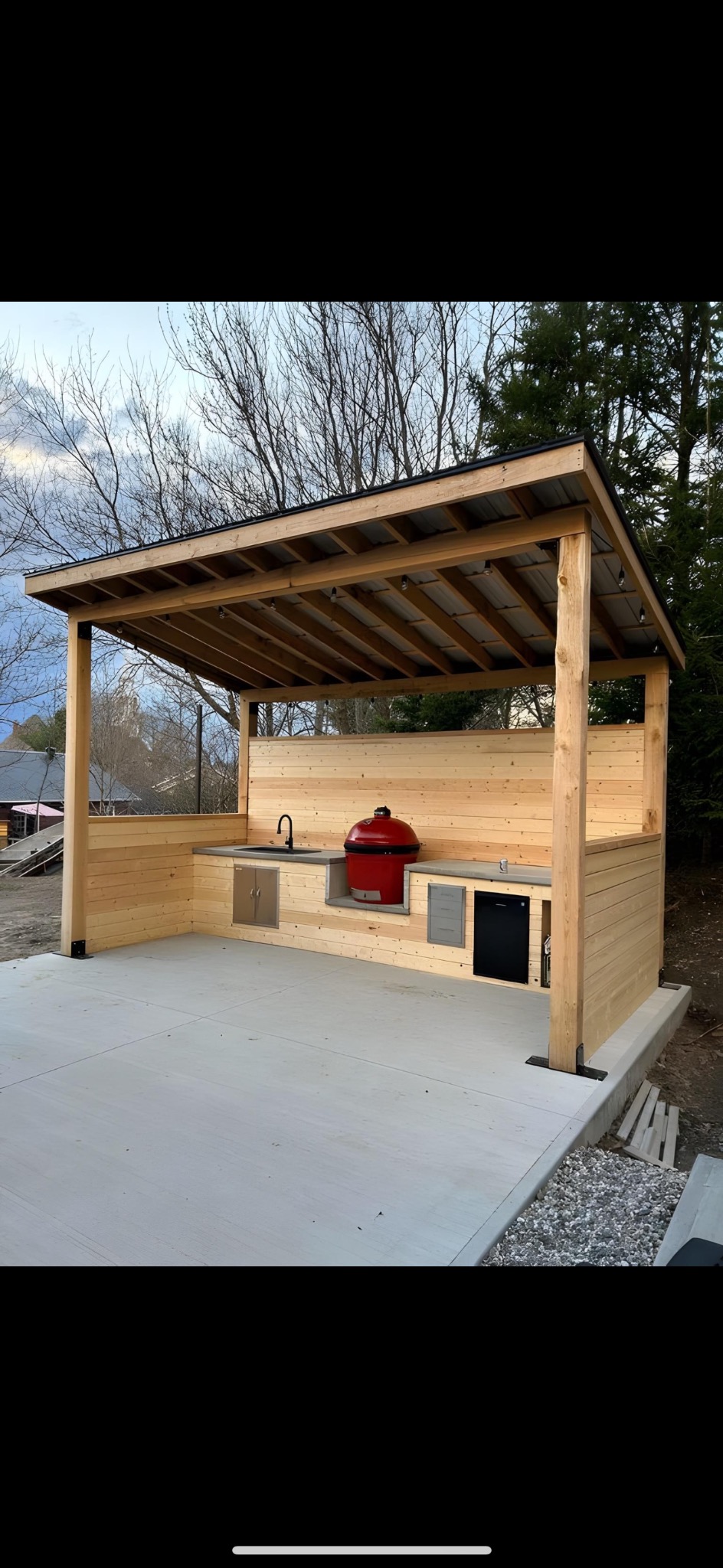 Zadaszona letnia kuchnia z drewnianą zabudową, wbudowanym grillem ceramicznym w kolorze czerwonym, zlewem i małą lodówką, oświetlona sznurem żarówek.