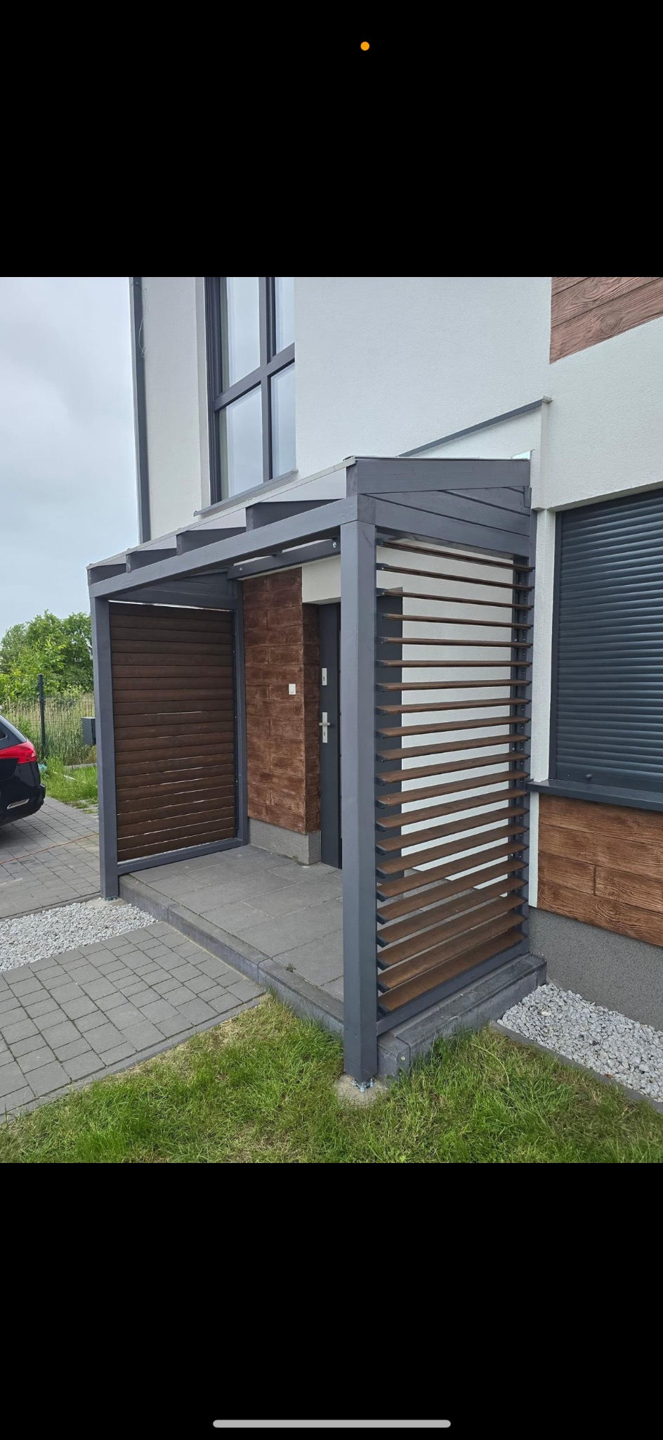 Nowoczesna pergola z szarymi ramami i drewnianymi listwami, przylegająca do budynku z elewacją imitującą cegłę, widoczna kostka brukowa i trawnik.