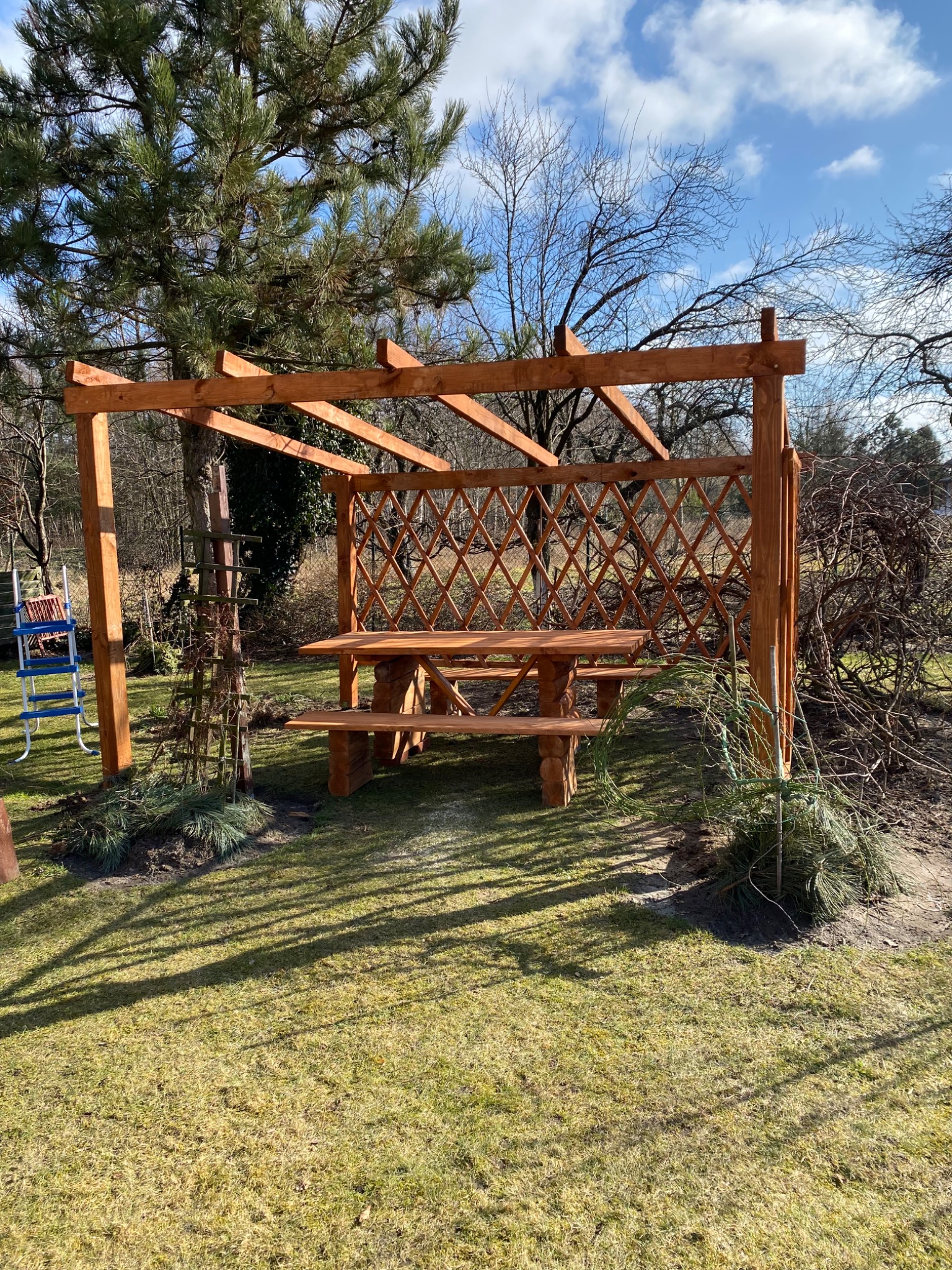 Drewniana pergola z ażurową ścianką i stołem z ławkami w ogrodzie, widoczna drabina malarska, w tle drzewa iglaste i liściaste.