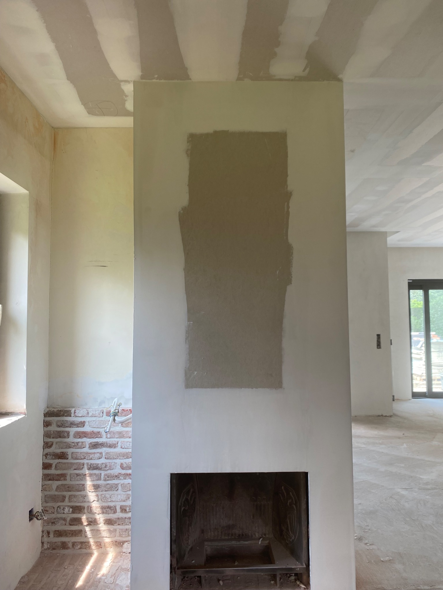 Wnętrze w trakcie prac wykończeniowych: częściowo wyszpachlowana ściana z otworem na kominek, fragment ściany z cegły oraz sufit z widocznymi pasami szpachli, podłoga zabezpieczona folią.