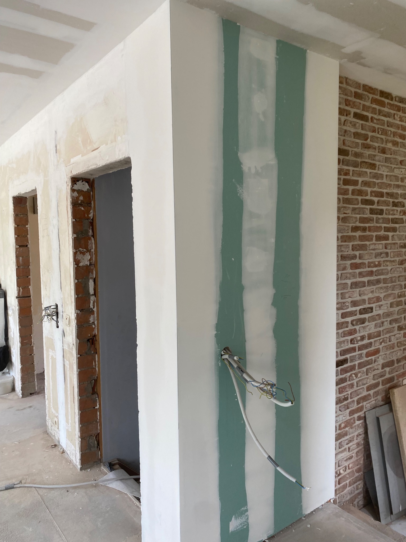 Wnętrze w trakcie prac wykończeniowych: ściana z częściowo położoną płytą gipsowo-kartonową z widocznymi przewodami elektrycznymi, obok fragment ściany z cegły i surowy otwór drzwiowy z odsłoniętą...