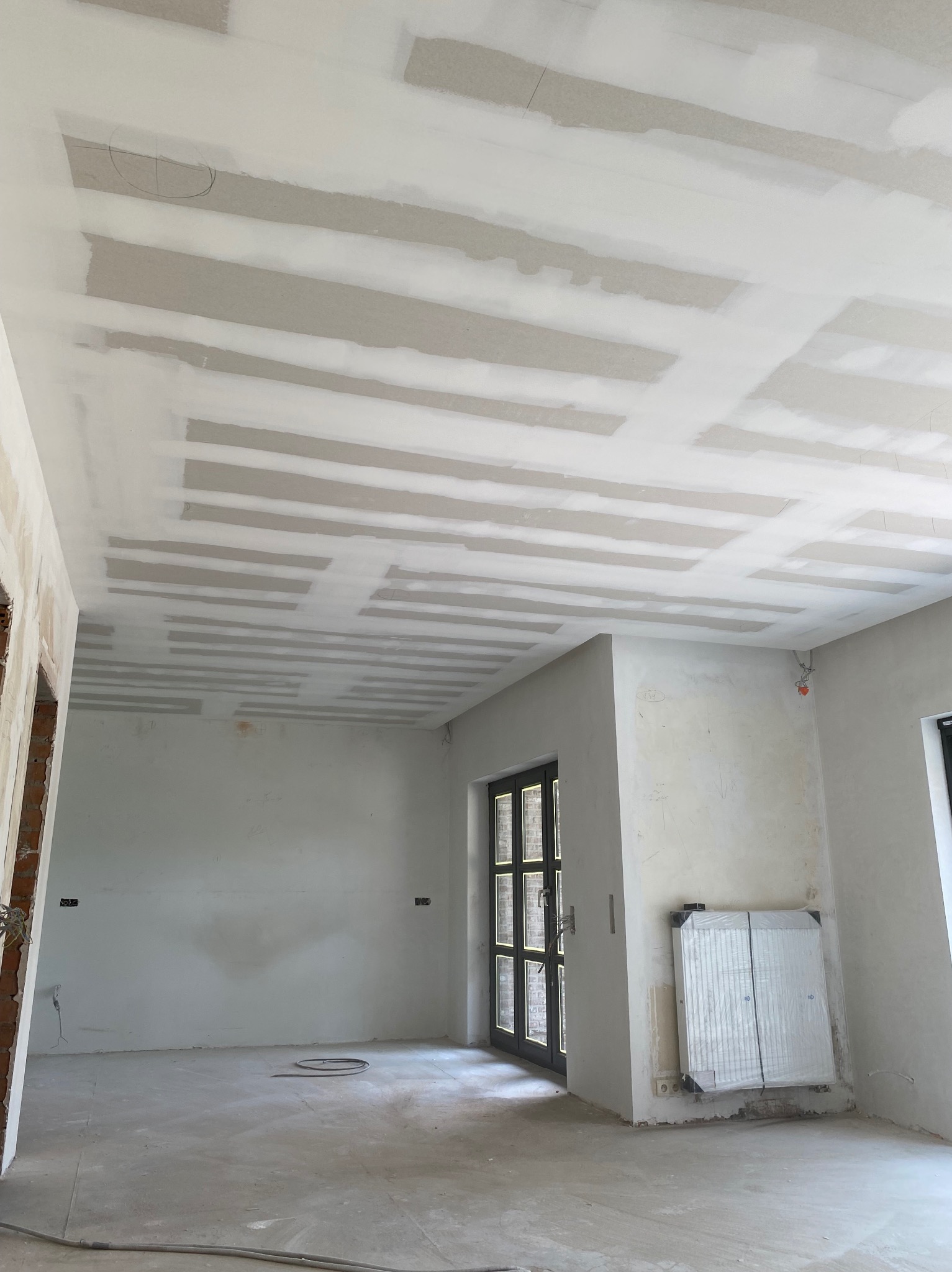 Wnętrze w trakcie remontu, widoczny sufit z zamontowaną konstrukcją pod płyty gipsowo-kartonowe, ściany przygotowane do dalszych prac wykończeniowych, okno z czarną ramą oraz grzejnik zabezpieczony...