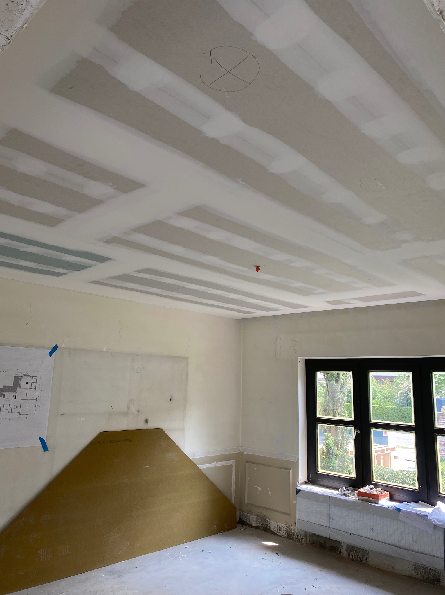 Montaż płyty karton gips na suficie w trakcie szpachlowania, widoczne łączenia płyt, rysunek techniczny na ścianie, okno z widokiem na zieleń.