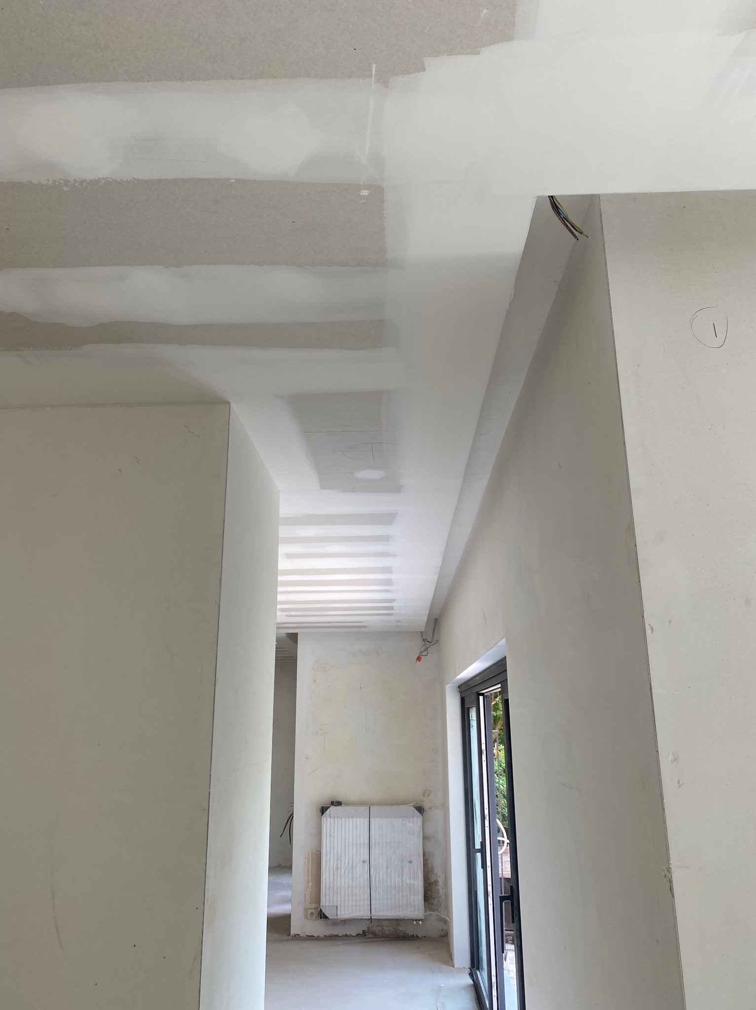 Wnętrze w trakcie prac wykończeniowych: sufit z płyt kartonowo-gipsowych ze szpachlowanymi łączeniami, widoczne przewody elektryczne, ściany w trakcie gruntowania, grzejnik w tle.