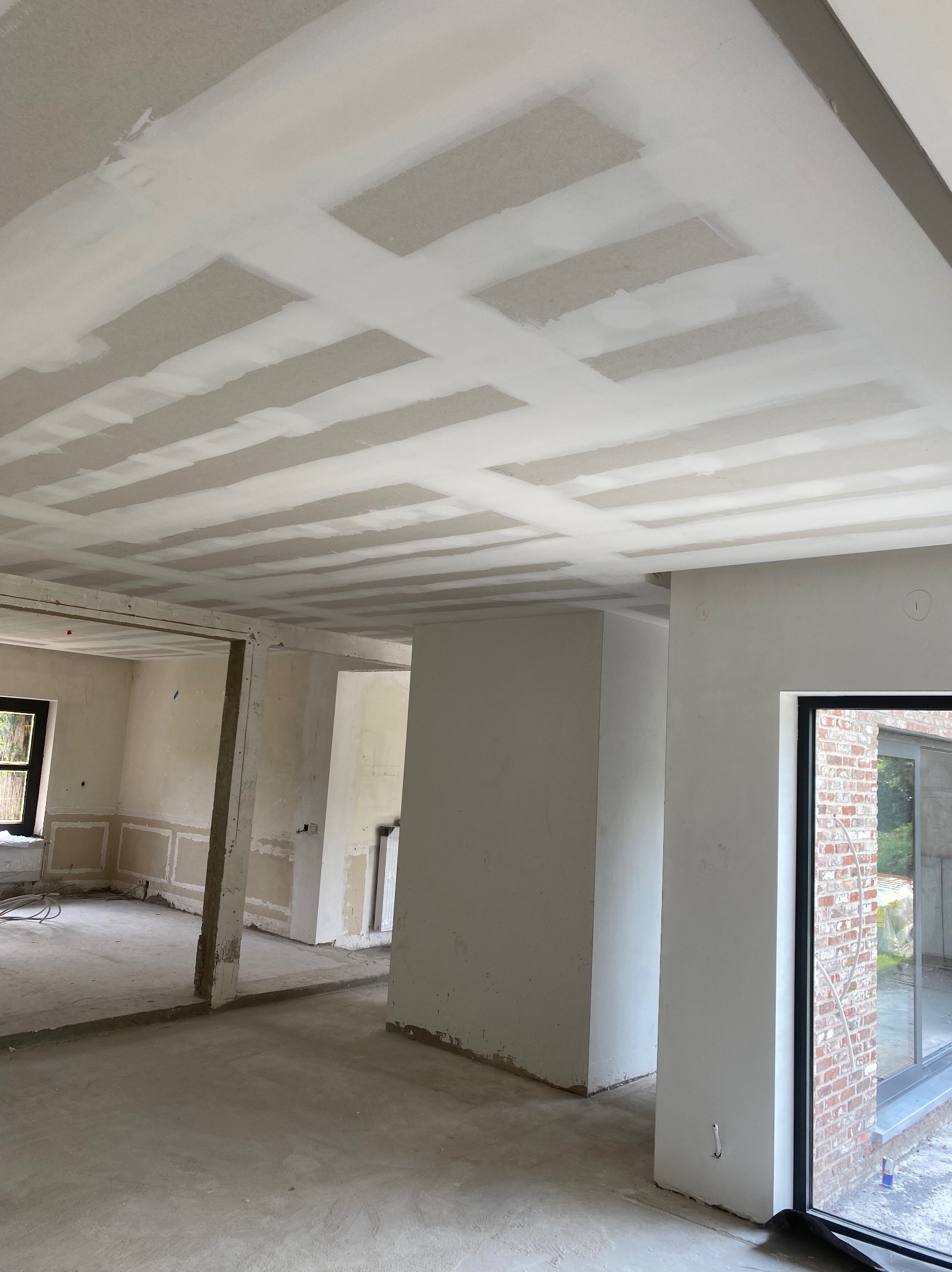Wnętrze w trakcie remontu: sufit z zamontowanymi płytami i częściowo zaszpachlowanymi łączeniami, widoczne przygotowane ściany i surowy beton na podłodze, duże okno z widokiem na ceglany mur.
