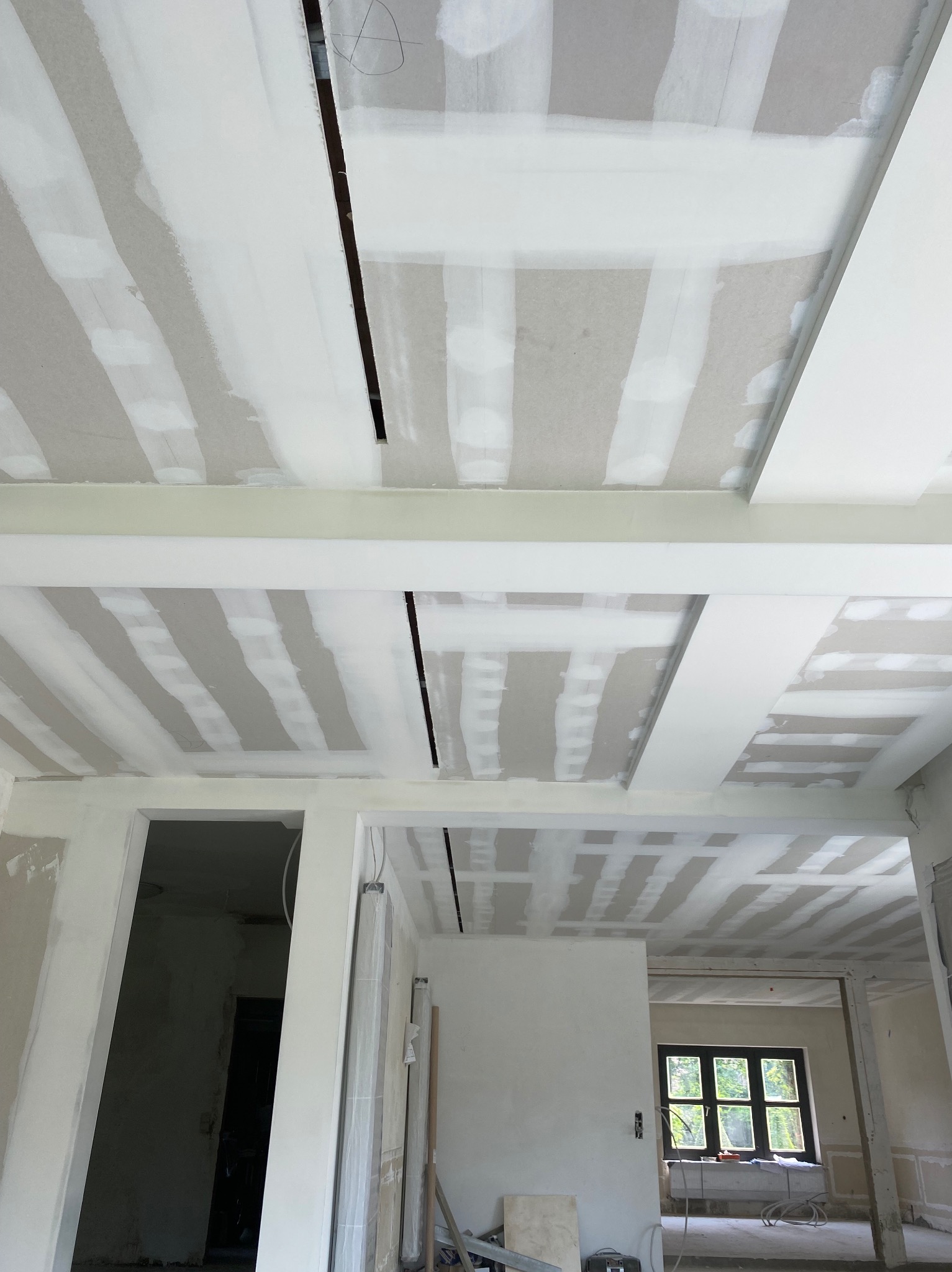 Sufit podwieszany z płyt kartonowo-gipsowych w trakcie montażu, widoczne łączenia maskowane taśmą, fragment instalacji elektrycznej w szczelinach, surowy stan wykończenia wnętrza.