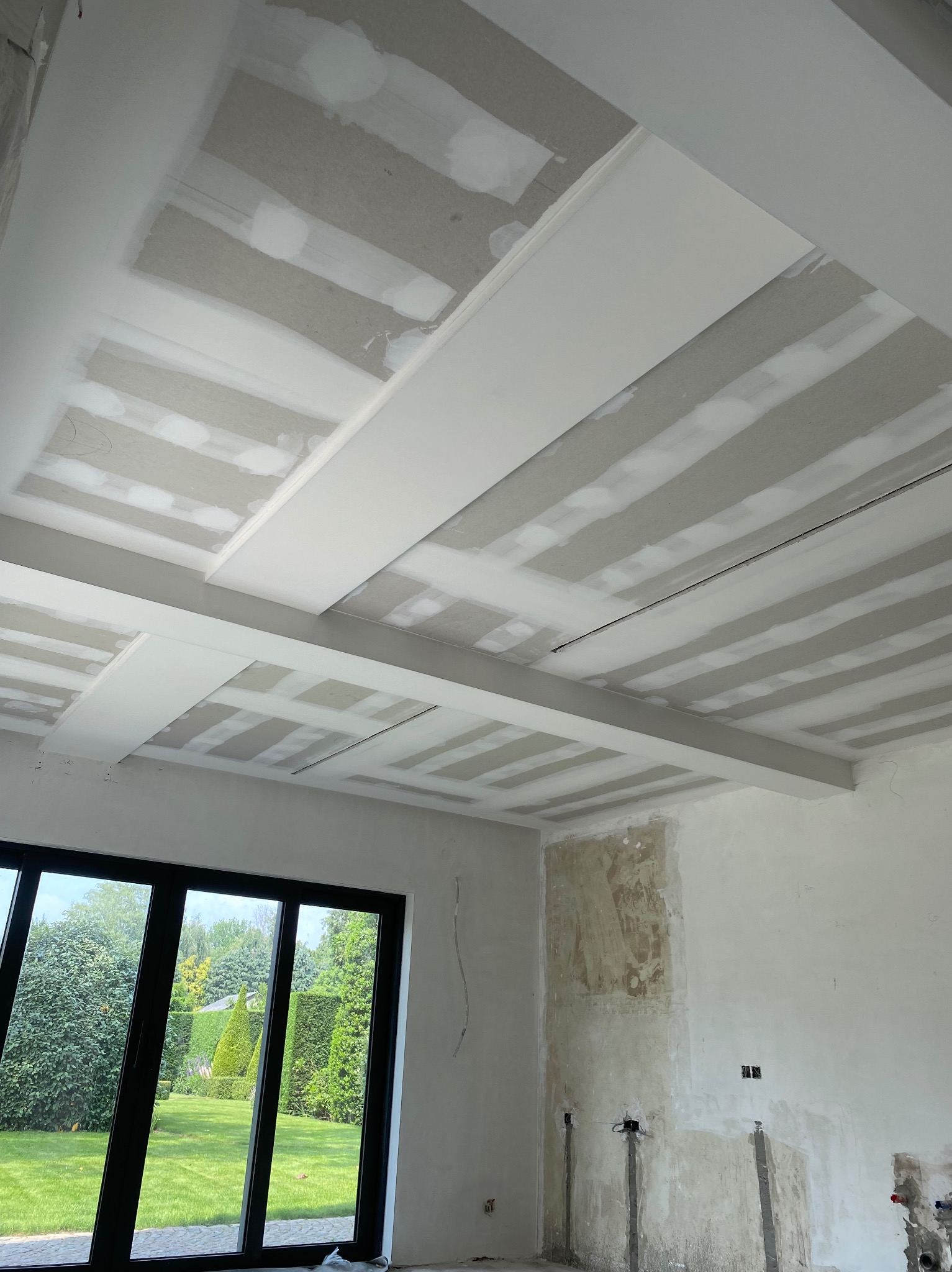 Sufit podwieszany ze szpachlowanymi łączeniami płyt w trakcie prac wykończeniowych w Zbytkowie, widok na duże okno z ogrodem w tle.