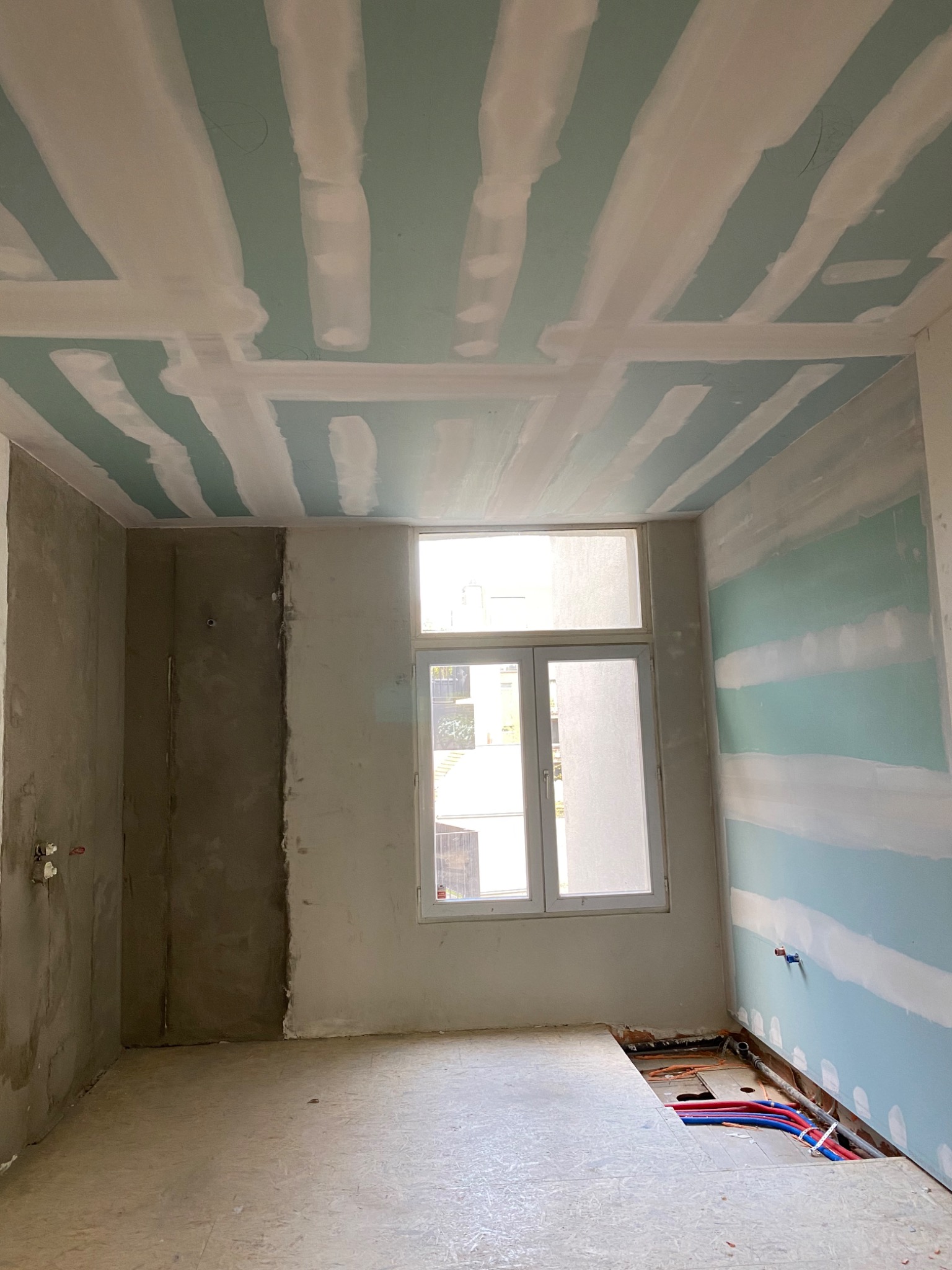 Wnętrze w trakcie remontu: ściany z nałożoną gładzią szpachlową, przygotowane do malowania, sufit z widocznymi łączeniami płyt gipsowo-kartonowych, odsłonięte instalacje wodne w podłodze.