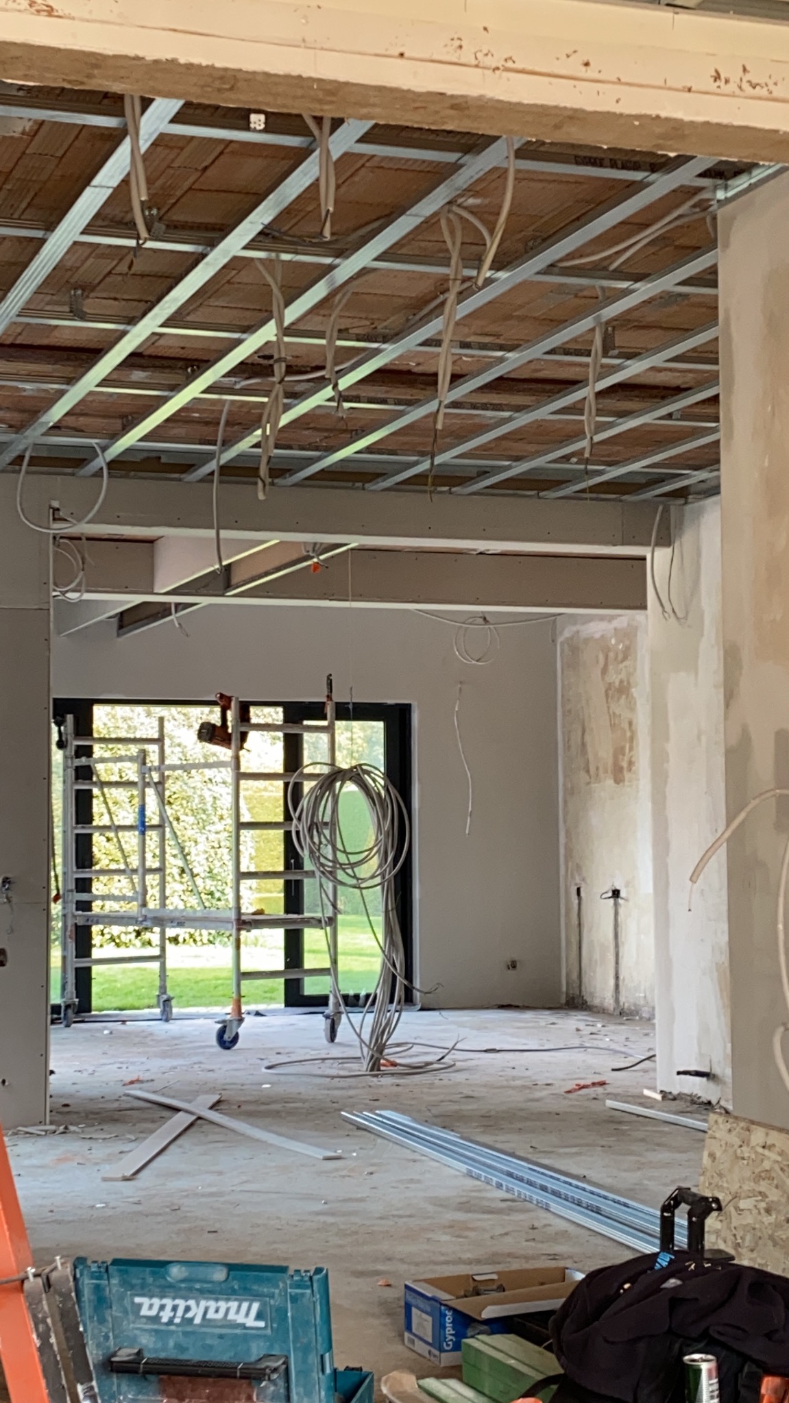 Wnętrze w trakcie remontu: widoczna konstrukcja sufitu podwieszanego z instalacją elektryczną, rusztowanie przy oknie z widokiem na ogród, narzędzia i materiały budowlane na podłodze.