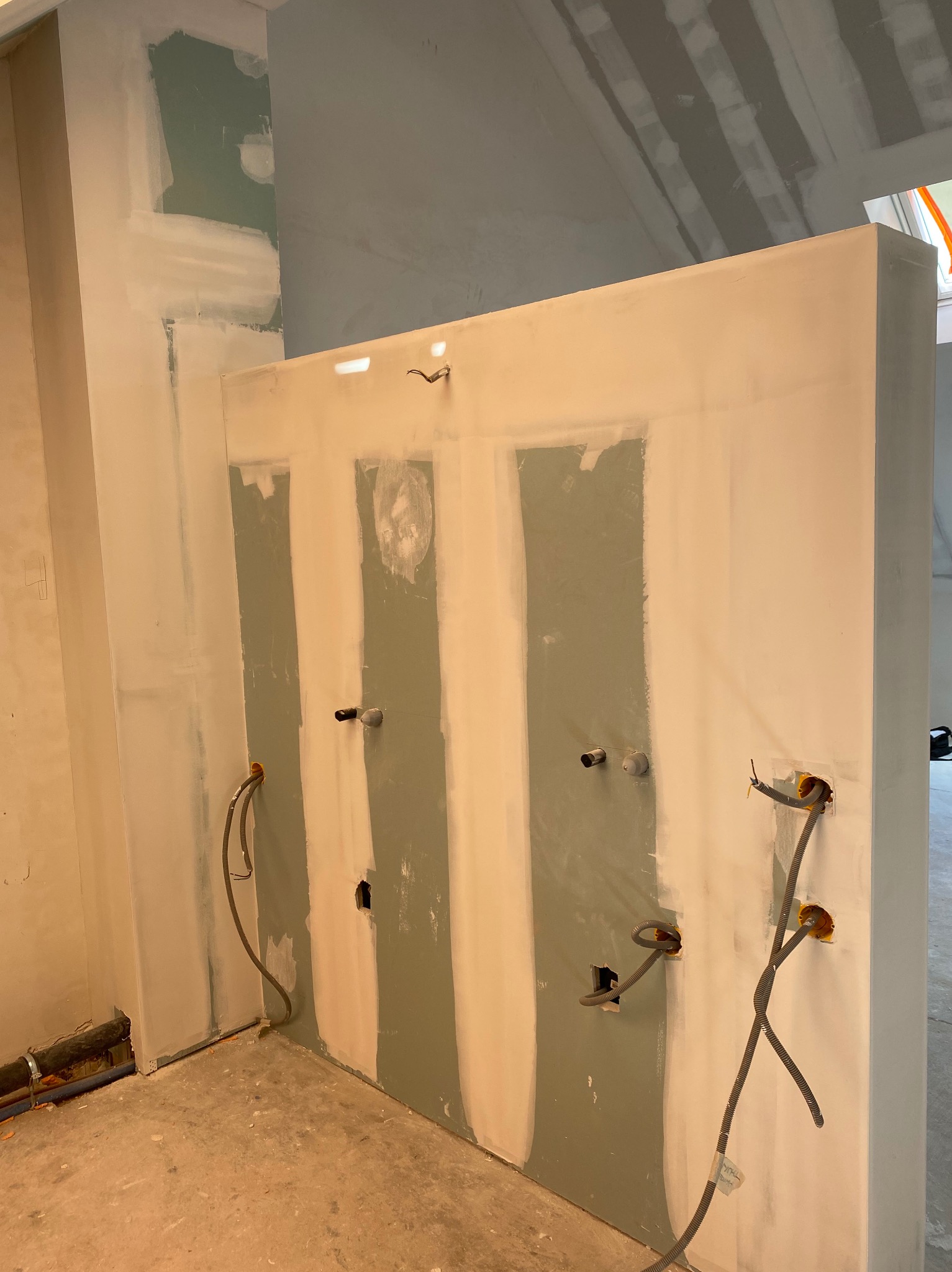 Ściana w trakcie szpachlowania w łazience, widoczne wystające kable i rury, ściany częściowo pokryte gładzią, surowy stan wykończenia.