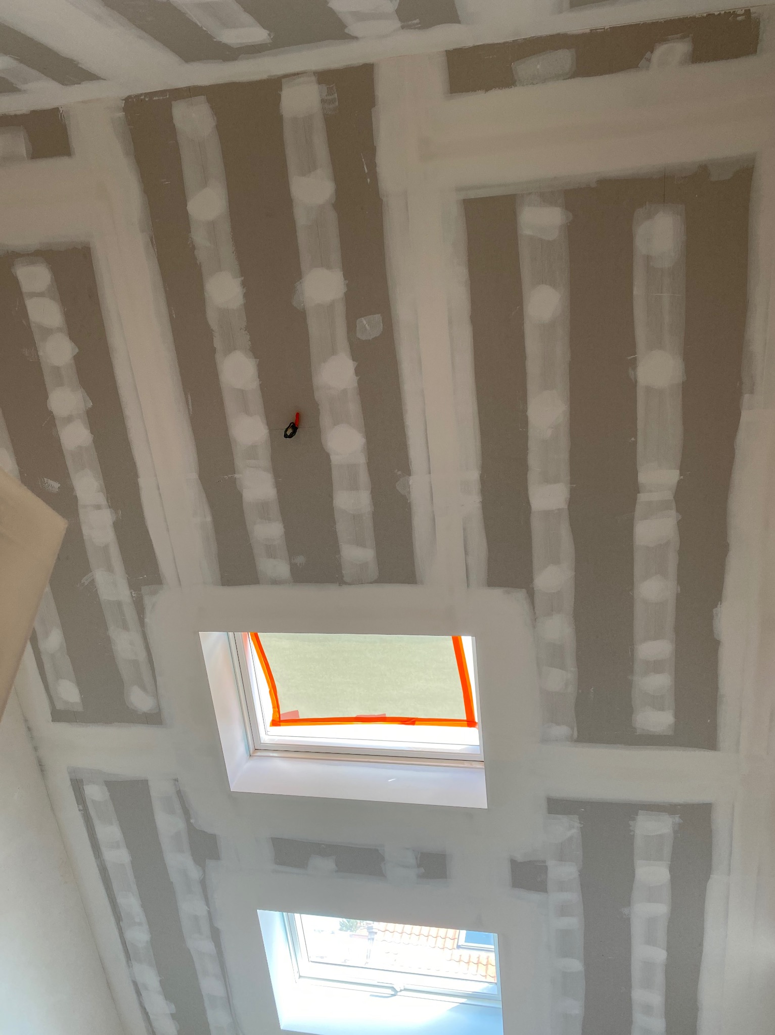 Sufit poddasza z oknami dachowymi w trakcie montażu płyt gipsowo-kartonowych, widoczne profile i miejsca łączeń szpachlowane na biało, zabezpieczona folią rama okna.