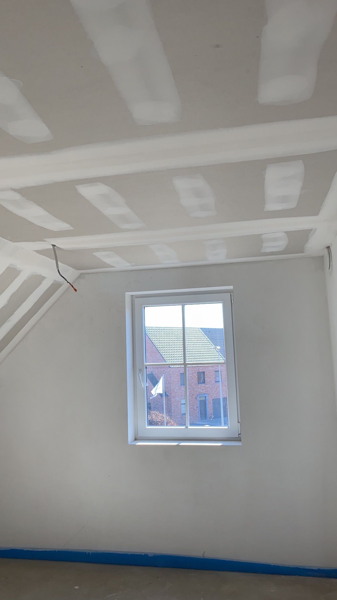 Pokój w trakcie remontu: widoczne płyty karton gips na suficie i ścianach, okno z widokiem na budynek z czerwonej cegły, wystające przewody elektryczne, niebieska taśma malarska zabezpieczająca...
