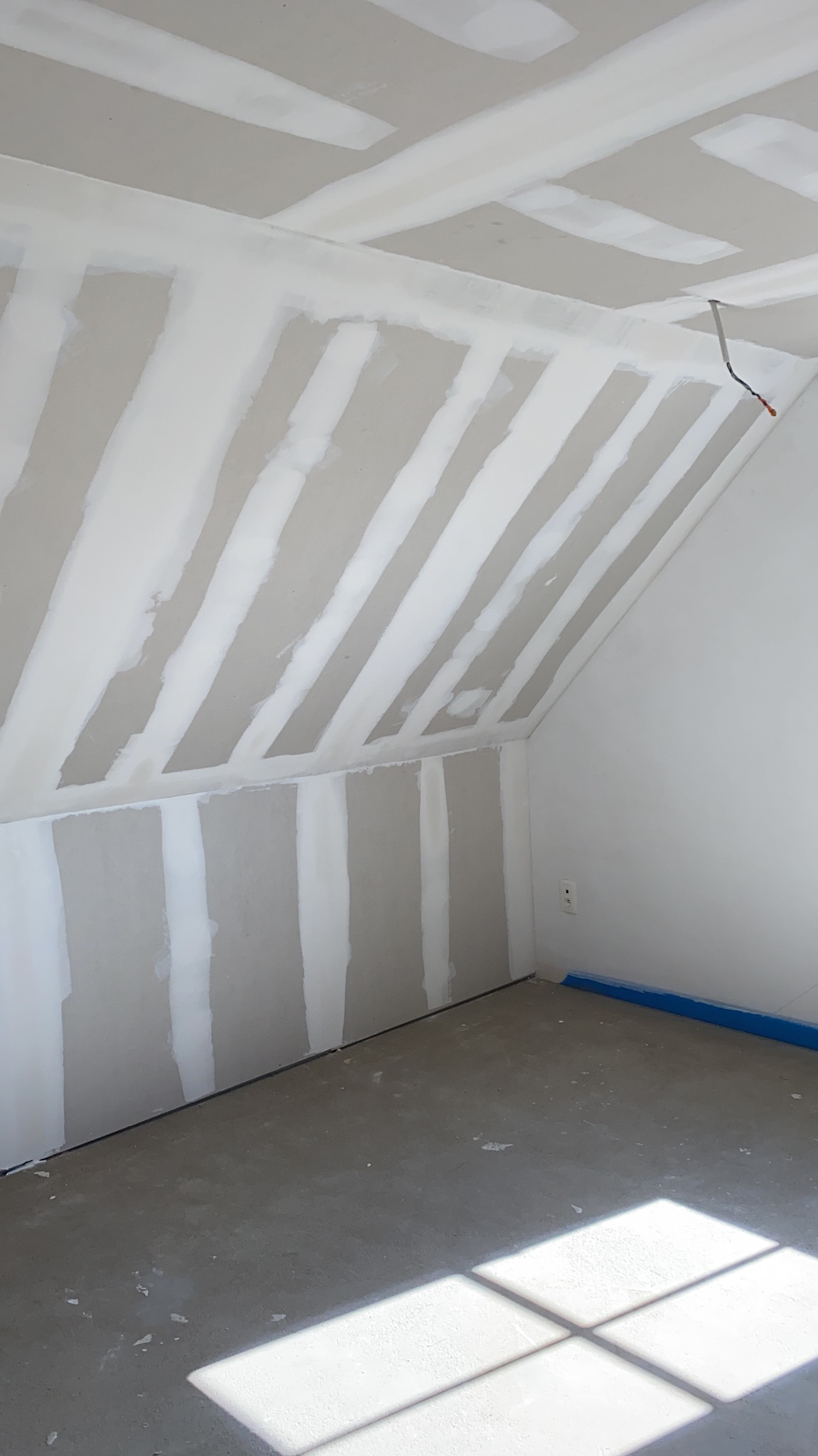 Poddasze w trakcie prac wykończeniowych: widoczne płyty gipsowo-kartonowe na skosach sufitu i ścianach, łączenia szpachlowane, surowa wylewka na podłodze, kabel zwisający z sufitu, okno dachowe...