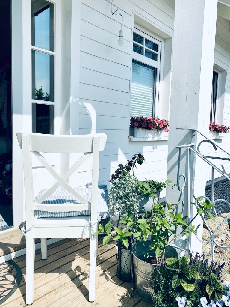 Jasny, drewniany taras w Nowym Sączu z białym krzesłem z poduszką w paski, otoczony różnorodnymi roślinami w donicach i wiadrach, z białymi ścianami domu w tle i kwiatami w oknach.