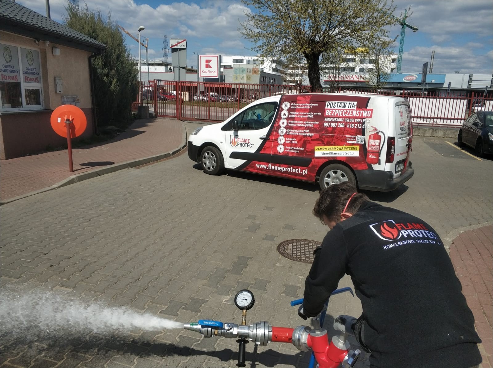 Testowanie instalacji hydrantowej w Warszawie przez pracownika w uniformie z logo firmy Flame Protect, na tle firmowego samochodu i budynku Kauflandu.