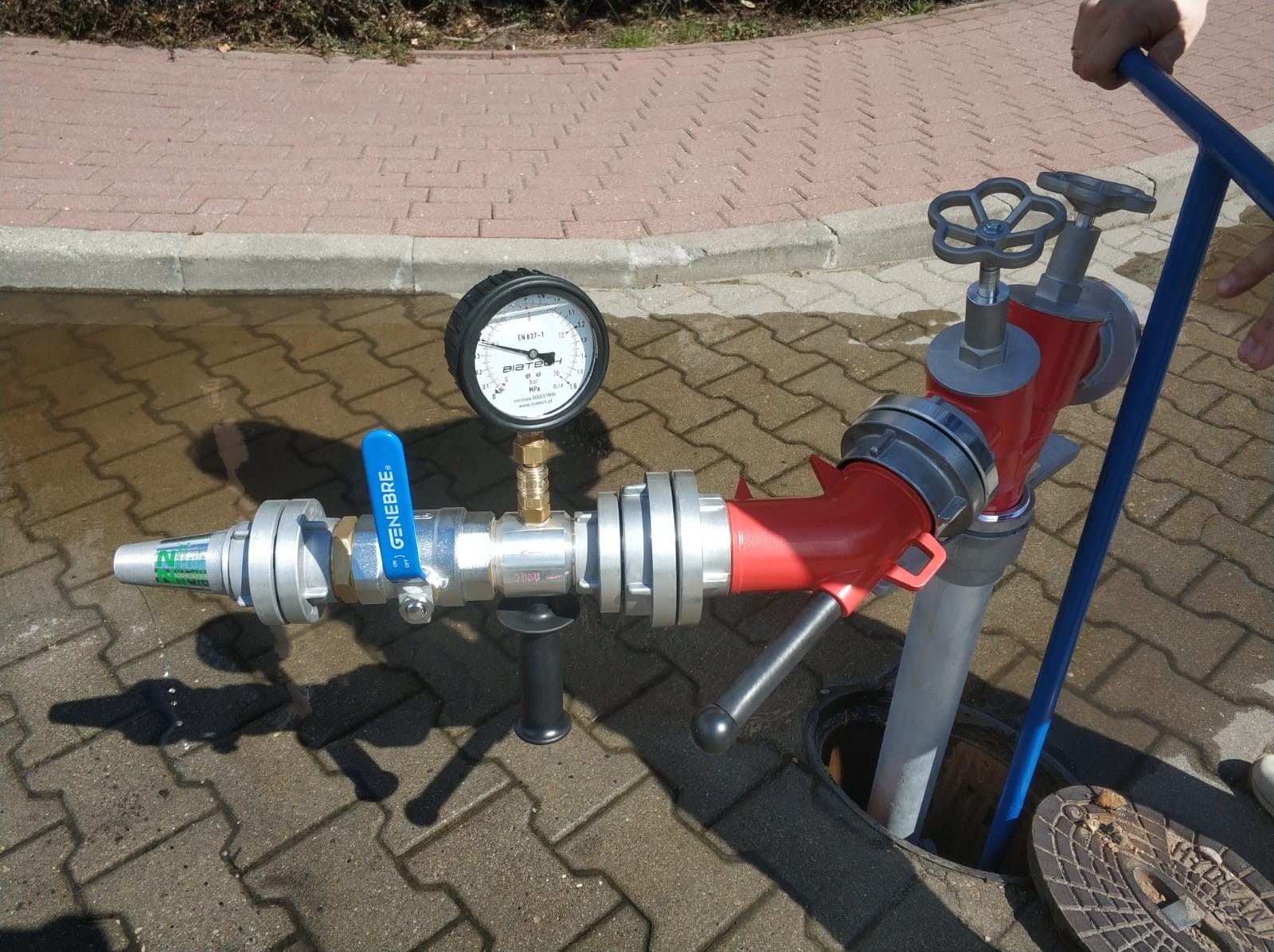 Czerwony hydrant pod ciśnieniem z manometrem, widoczny na kostce brukowej, z otwartą studzienką i fragmentem buta w tle.