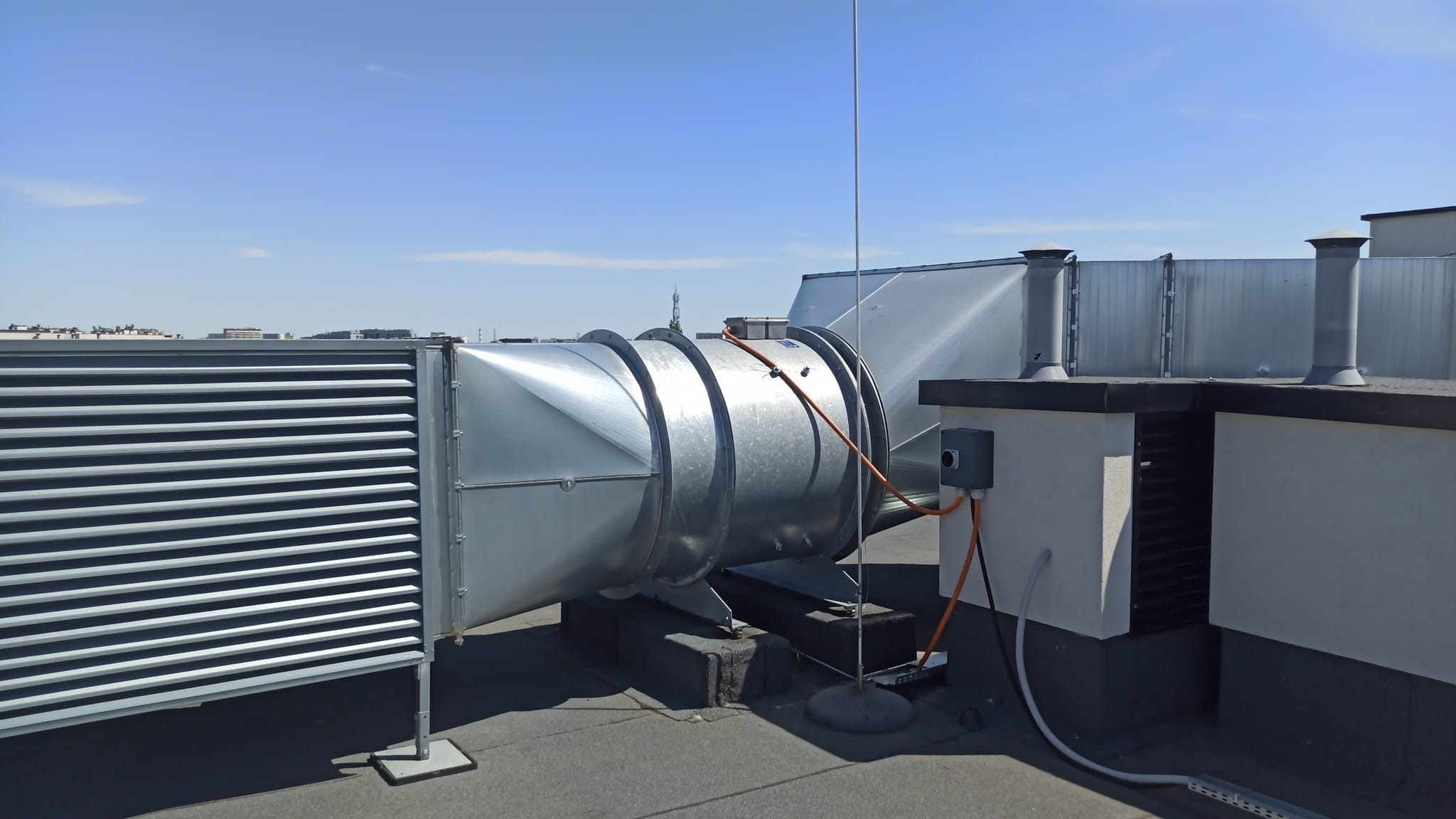 System wentylacyjny z metalowymi rurami i kratkami na dachu budynku, widoczne przewody elektryczne i antena odgromowa.