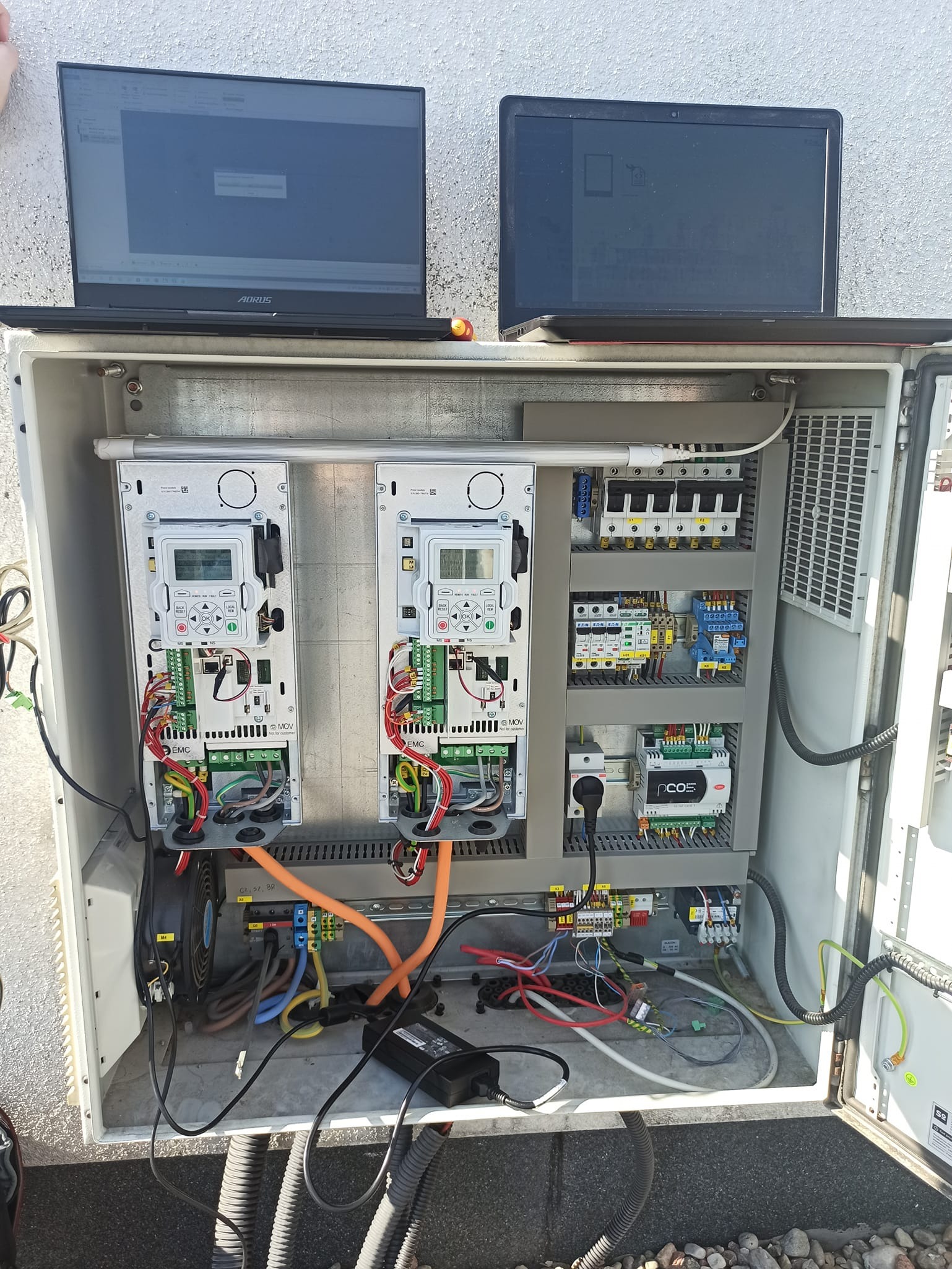 Otwarta szafa sterownicza z dwoma panelami kontrolnymi, okablowaniem i elementami elektrycznymi, z dwoma laptopami na górze, jeden marki Aorus, sugerującymi konfigurację lub diagnostykę systemu.