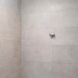 Ściana łazienki wyłożona jasnoszarymi, prostokątnymi płytkami ceramicznymi, z fragmentem ściany obłożonej mozaiką imitującą drewno oraz chromowanym uchwytem na słuchawkę prysznicową.