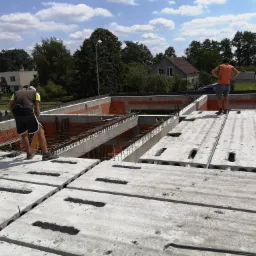 Budowa domu: Robotnicy na stropie Teriva z widocznymi pustakami, zbrojenie na obrzeżach, w tle zabudowa jednorodzinna i drzewa.