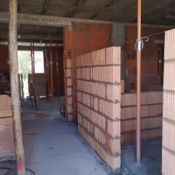 Wnętrze budynku w trakcie budowy w Opalenicy, widoczne ściany z czerwonej cegły ceramicznej, podparte metalowymi stemplami, surowy betonowy strop.