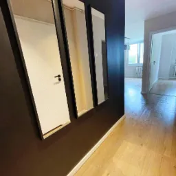 Korytarz z trzema pionowymi lustrami w czarnych ramach na ciemnej ścianie, odbijającymi białe drzwi z geometrycznym wzorem, widok na jasne pomieszczenie z grzejnikiem i oknem.