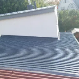 Nowy, ciemnoszary dach z blachy trapezowej na budynku z białą elewacją, widoczny fragment starego, czerwonego dachu i otoczenie z zielenią.