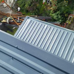 Widok z góry na fragment szarego dachu z blachy trapezowej, z widocznymi śrubami mocującymi, w tle ogród z drewnianym płotem i stertą drewna.