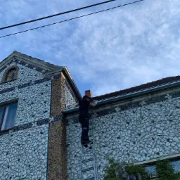 Pracownik na drabinie przy domu z elewacją z potłuczonych talerzy, montujący lub naprawiający rynnę pod okapem dachu.