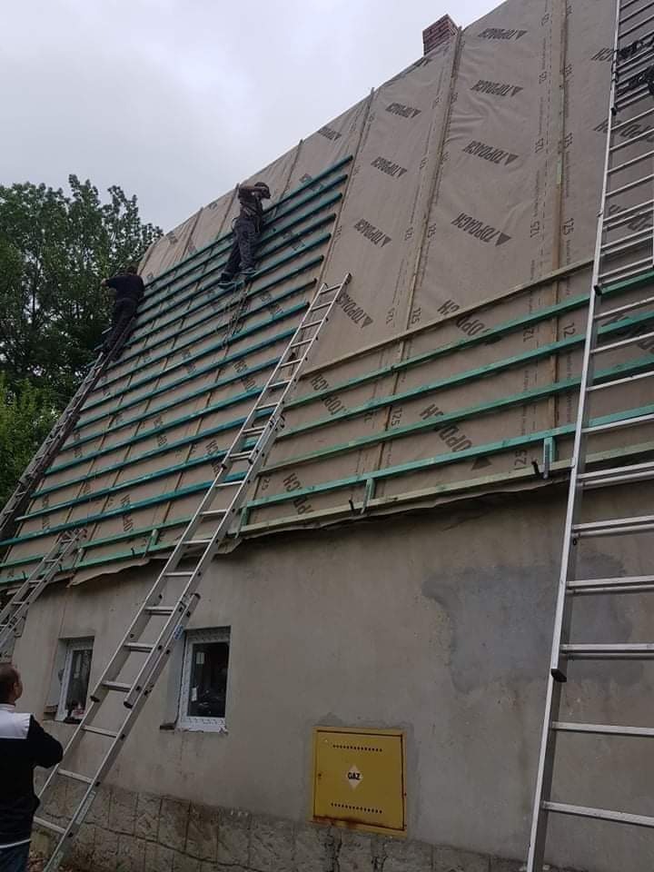 Montaż łat dachowych na folii wierzchniej dachu domu jednorodzinnego, widoczne drabiny i pracownicy na dachu.