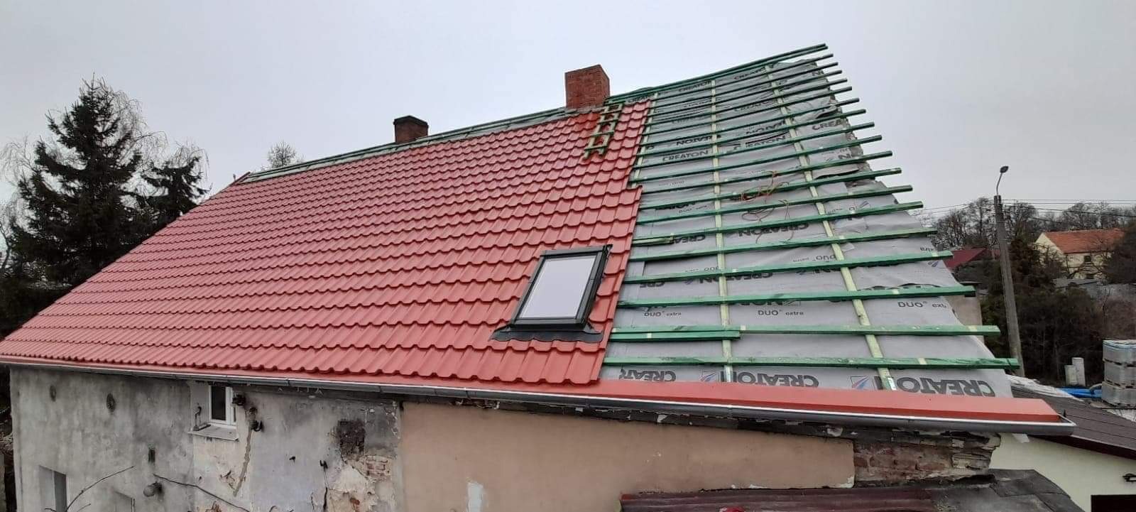 Częściowo pokryty czerwonymi dachówkami dach domu jednorodzinnego z oknem dachowym, widoczna konstrukcja z łat i membrany na pozostałej części dachu.