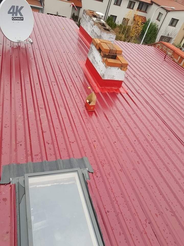 Czerwony dach z blachy trapezowej po deszczu, widoczne dwa kominy z cegły i betonu, okno dachowe z szarą obróbką oraz antena satelitarna 4K.